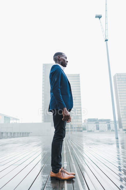 Elegante uomo d'affari in piedi sul marciapiede sotto la pioggia contro i moderni edifici della città — Foto stock