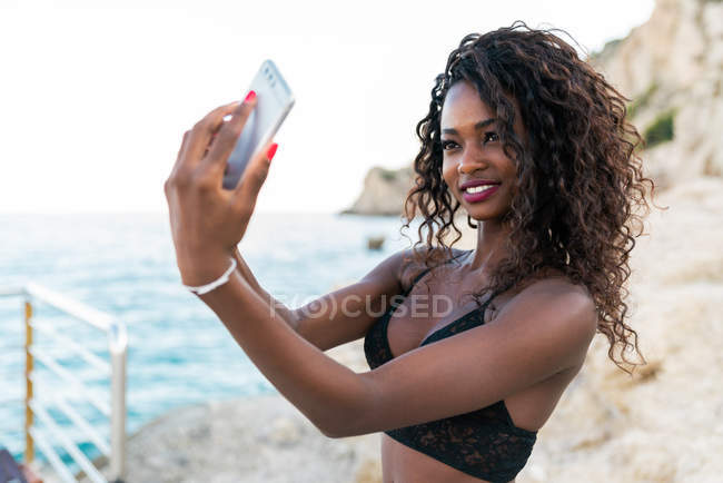 Cheerful pretty black woman taking selfie with mobile phone on coast — Stock Photo