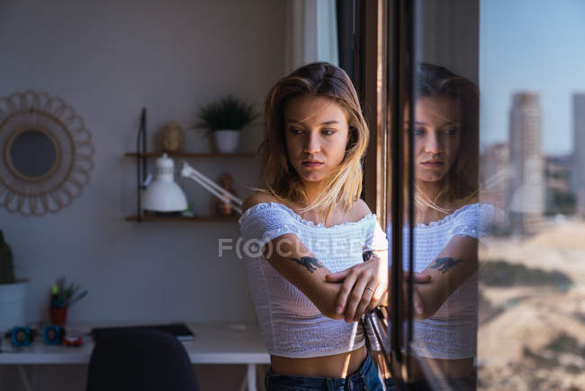 Mujer bastante pensativa de pie cerca de la ventana y mirando hacia otro lado - foto de stock
