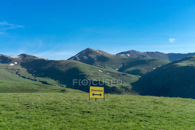 Green landscape of mountains and yellow sign showing diversion with arrows in different directions under blue sky — Stock Photo