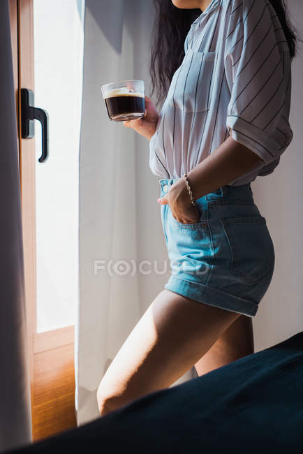 Безликая женщина в джинсовых шортах и рубашке, стоящая у окна со стеклянной чашкой кофе — стоковое фото