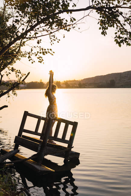 Woman in dress with hands up standing on sunken wooden pier on lake at sunset — Stock Photo