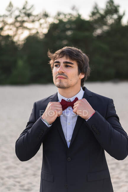 Adult man wearing elegant black suit with bow tie and standing on beach with hands in pockets looking at camera — Stock Photo