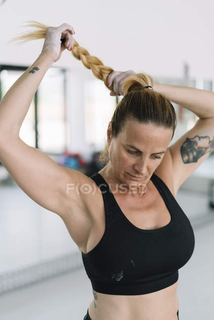 Strong blonde woman in sports bra making braid while training in gym — Stock Photo