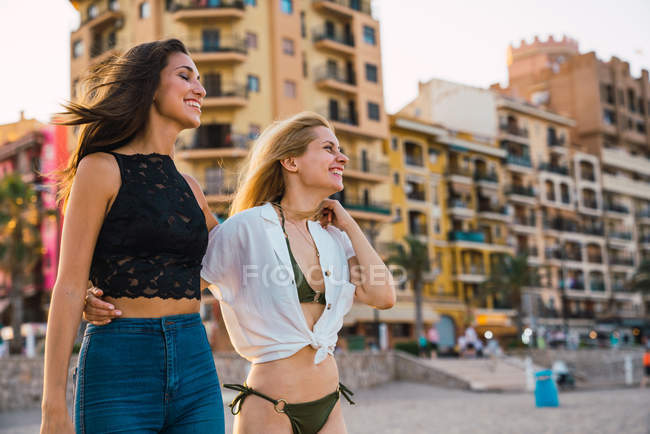 Laughing female friends walking on beach with buildings on background — Stock Photo