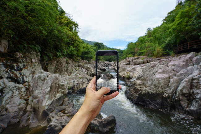 Turista de cultivos usando smartphone y tomando fotos del arroyo de agua entre las rocas en Yanoda Rainforest, China - foto de stock