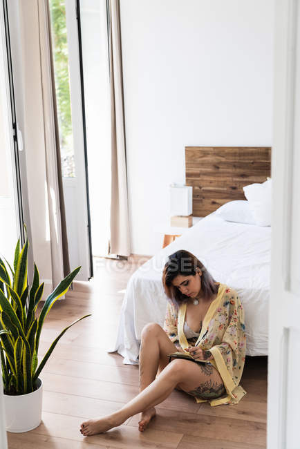 Jovem mulher em roupão de seda sentado no chão e desenho em caderno de esboços no quarto elegante — Fotografia de Stock