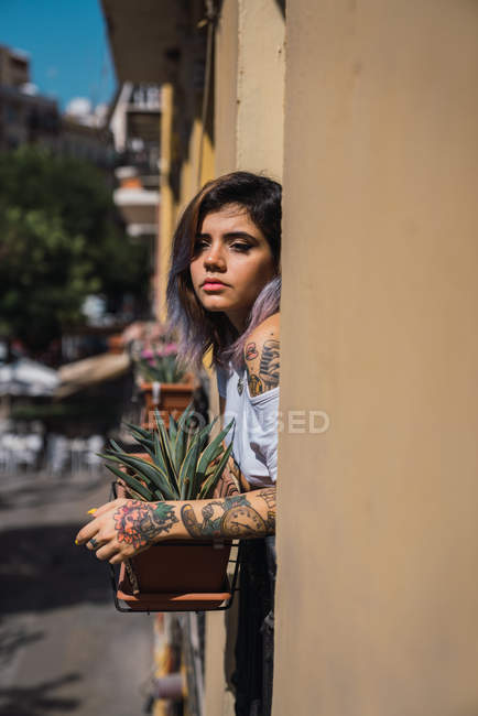 Jovem pensativa com tatuagens apoiando-se no corrimão da varanda e desfrutando da luz solar — Fotografia de Stock