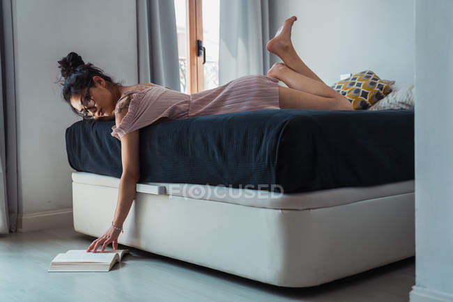 Focused sensual woman with eyeglasses lying on bed and reading book on floor — Stock Photo