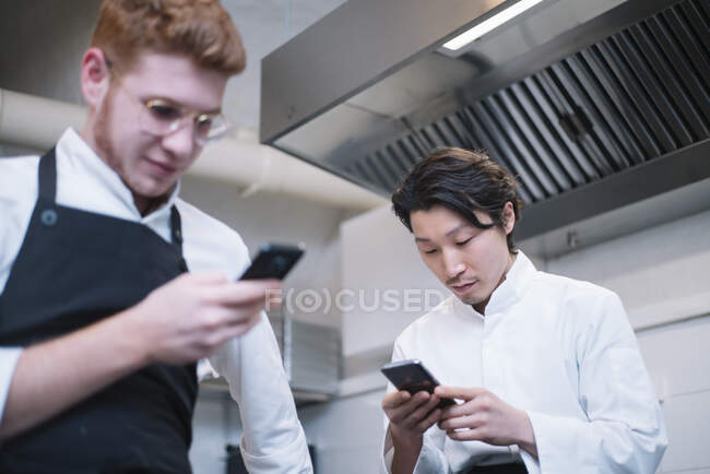 A partir de baixo tiro de dois caras em uniforme cozinheiro de pé na cozinha do restaurante e navegando smartphones durante o intervalo — Fotografia de Stock