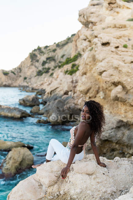 Elegant woman sitting on cliff above sea water and looking away — Stock Photo