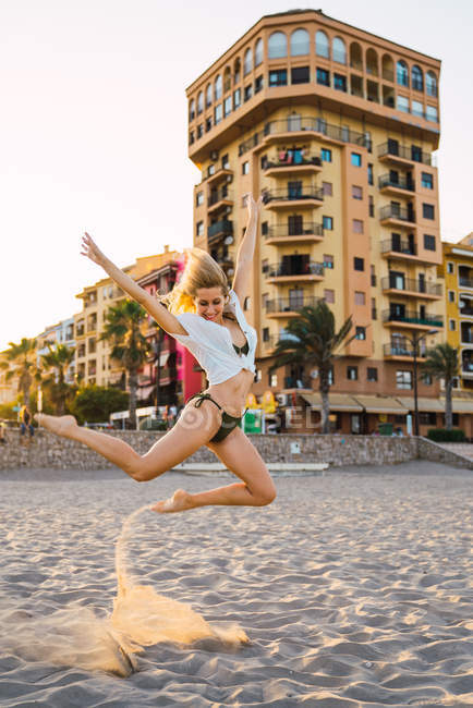 Гнучка молода жінка стрибає на пляжі з будівлями на фоні — стокове фото