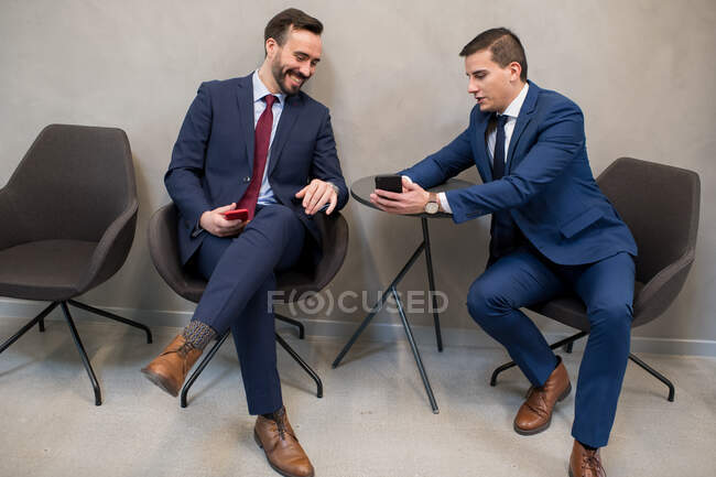 Два элегантных человека в костюмах и сидят на стульях в офисе, обмениваясь смартфонами и развлекаясь. — стоковое фото
