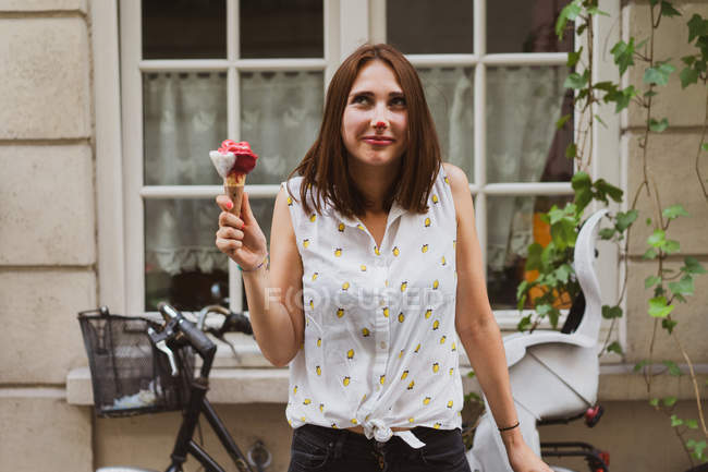 Улыбающаяся задумчивая женщина держит мороженое на улице напротив дома — стоковое фото