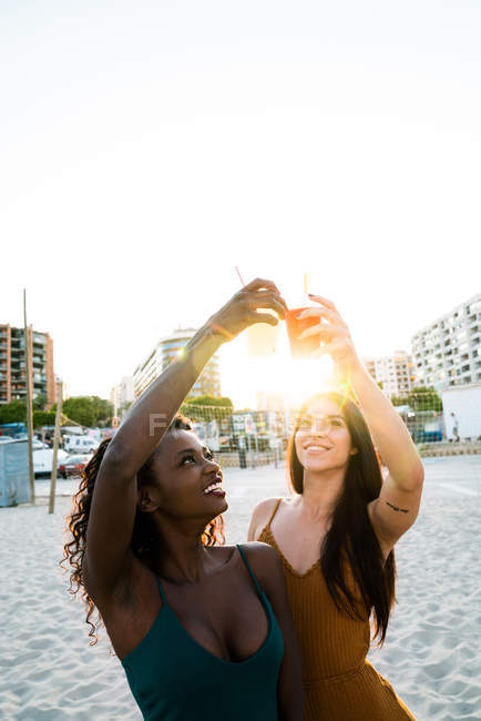 Stylische multiethnische Frauen klirren mit Tassen am Strand im Sonnenlicht — Stockfoto