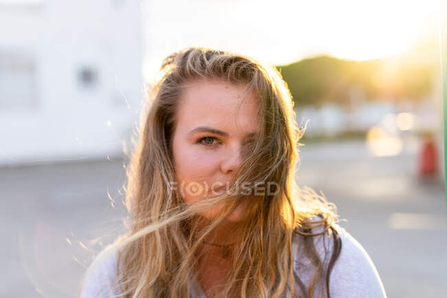 Mujer joven feliz en traje casual mirando a la cámara y riendo en la espalda iluminada por la luz del atardecer - foto de stock