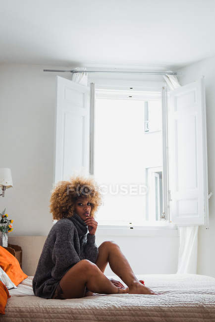 Етнічна жінка в сірому светрі сидить на ліжку вдома і дивиться на камеру — стокове фото