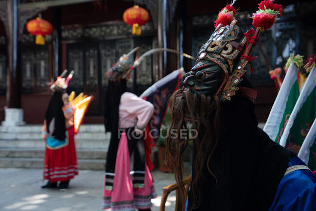 GUINZHOU, CHINA - JUNHO 14, 2018: Grupo de minorias étnicas Miao fêmeas em trajes tradicionais brilhantes e máscaras em pé com o homem em traje amarelo ao ar livre na parede do edifício chinês em Guizhou — Fotografia de Stock