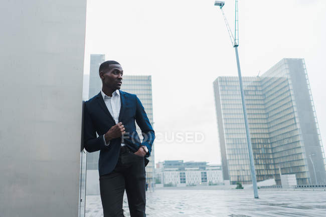 Afrikanisch-amerikanischer Geschäftsmann lehnt an einer Mauer im Freien mit modernen Gebäuden im Hintergrund — Stockfoto
