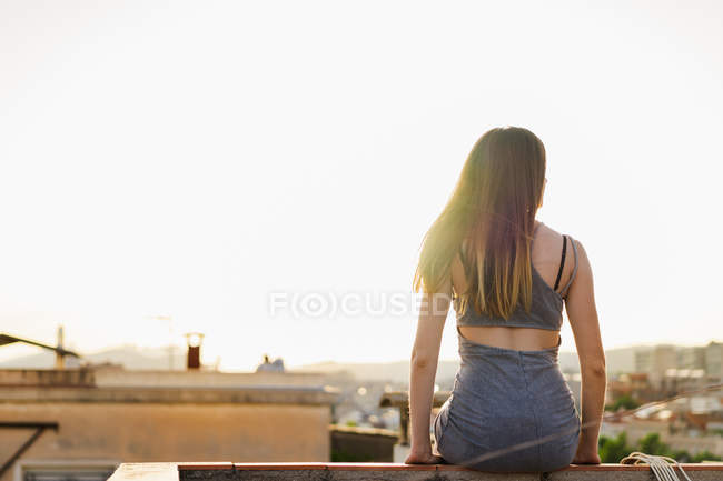 Brunette woman in summer dress sitting on parapet of roof with blurred town on background — Stock Photo
