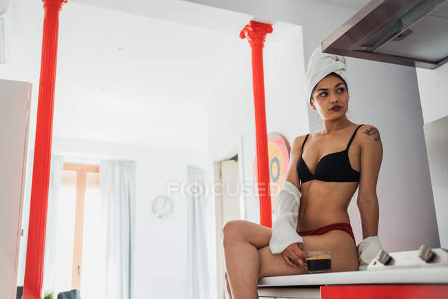 Donna sensuale in lingerie con asciugamano sulla testa seduta sul bancone con tazza di caffè — Foto stock