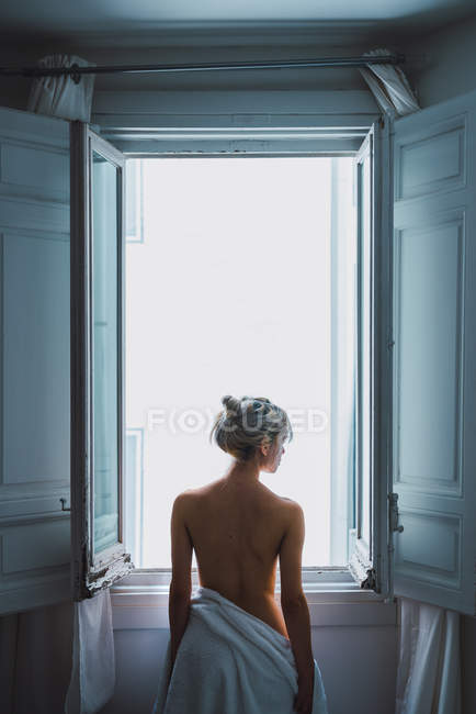 Vista trasera de mujer rubia desnuda con toalla blanca de pie cerca de la ventana abierta después de la ducha - foto de stock