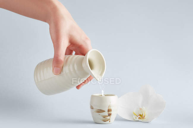 Human hand holding white ceramic pitcher and pouring water into cup with floral ornament on white background with white orchid — Stock Photo
