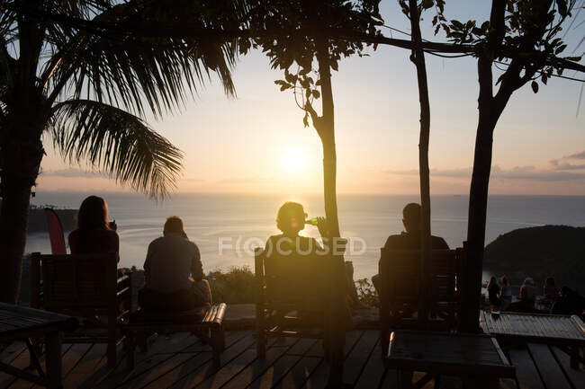 Vue arrière du groupe d'amis se détendre au bord de la mer dans les lumières du coucher du soleil en Thaïlande. — Photo de stock
