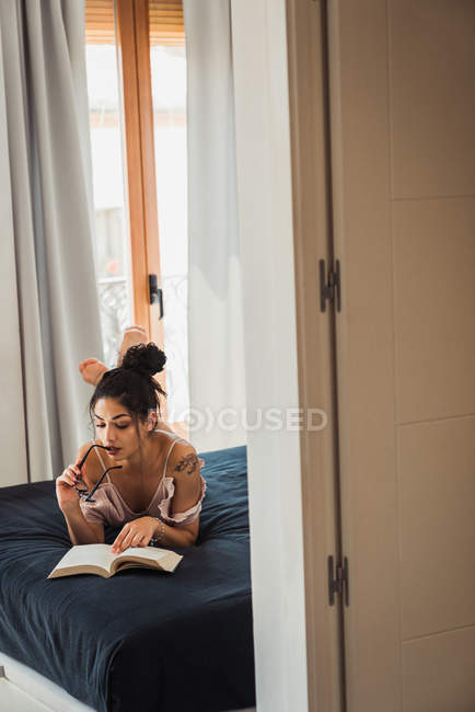 Очаровательная молодая женщина, кусающая очки, лежа на кровати и читая книгу — стоковое фото