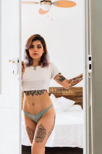 Молода татуйована жінка в трусиках і футболці дивиться на камеру, стоячи в дверях затишної спальні — стокове фото