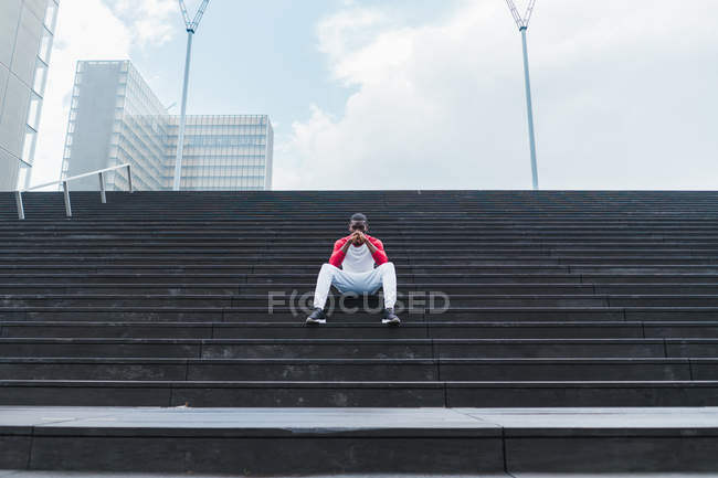 Homme ethnique en vêtements de sport assis sur des escaliers gris humides et la tête d'appui avec les mains pliées et regardant la caméra — Photo de stock