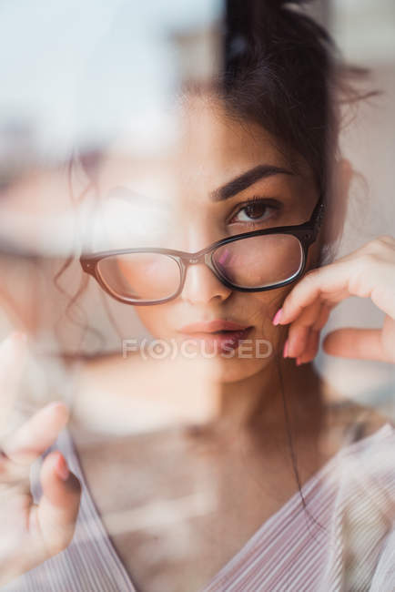 Femme sensuelle avec des lunettes regardant la caméra derrière la fenêtre — Photo de stock