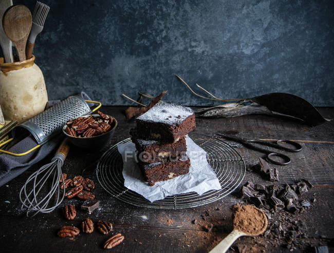 Кусочки вкусного шоколадного брауни на проволочной стойке с ингредиентами на темной деревянной поверхности — стоковое фото