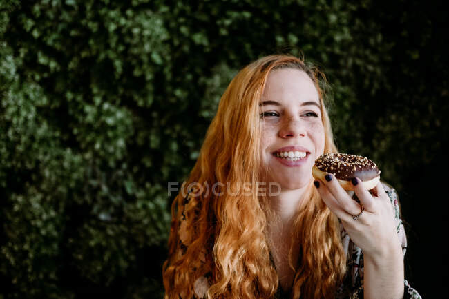 Cheerful redhead pretty woman with cup sitting at bush and looking away. — Stock Photo