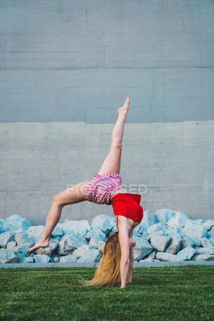 Jovem mulher fazendo suporte de mão na grama contra parede de concreto e pedras — Fotografia de Stock