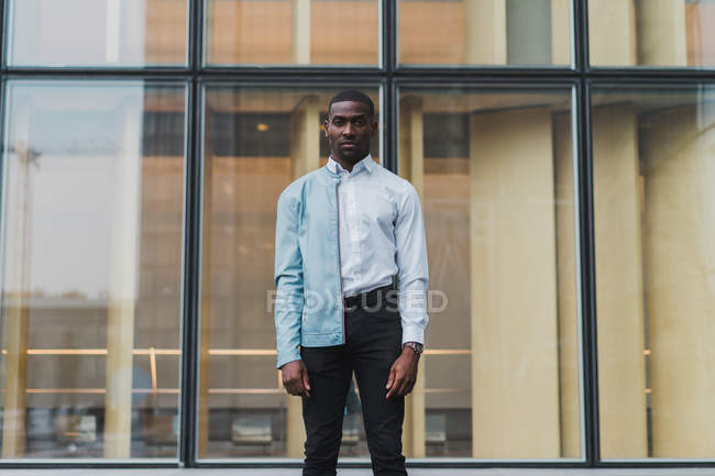Serious young ethnic man in white shirt and light leather jacket on one shoulder standing against glass modern building — Stock Photo