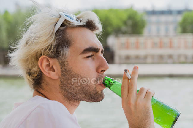 Blond bearded male holding cigarette in hand while drinking beer on street on blurred background — Stock Photo