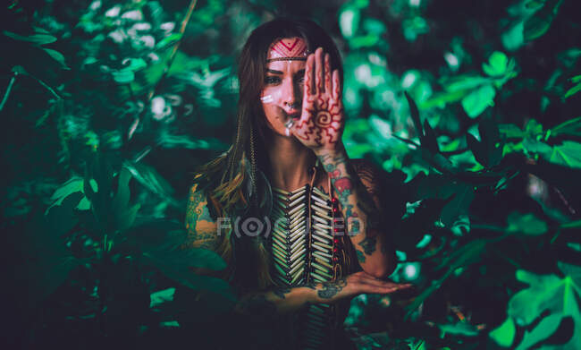Atractiva joven con pinturas indias tradicionales en la cara mirando a la cámara y de pie en el bosque - foto de stock