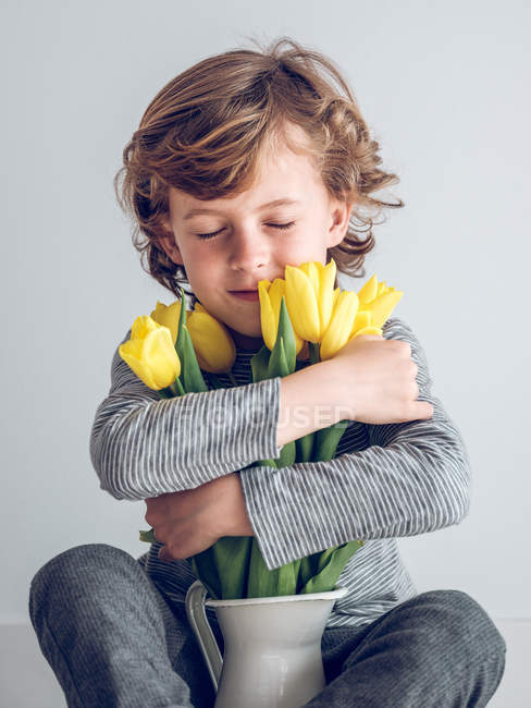 Молодий хлопчик з закритими очима сидить і приймає купу жовтих тюльпанів на сірому фоні — стокове фото