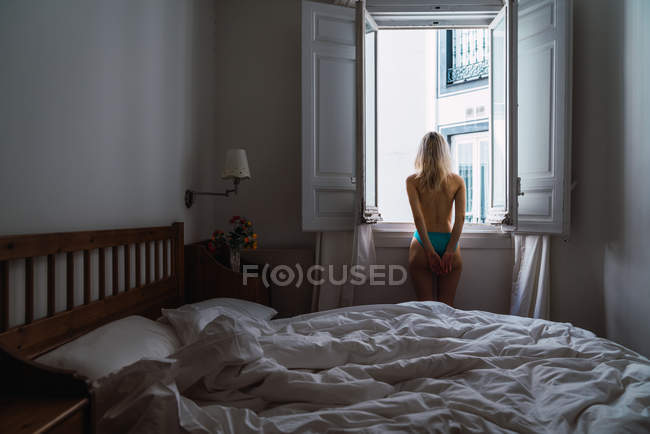 Без рубашки блондинка смотрит в окно в спальне — стоковое фото