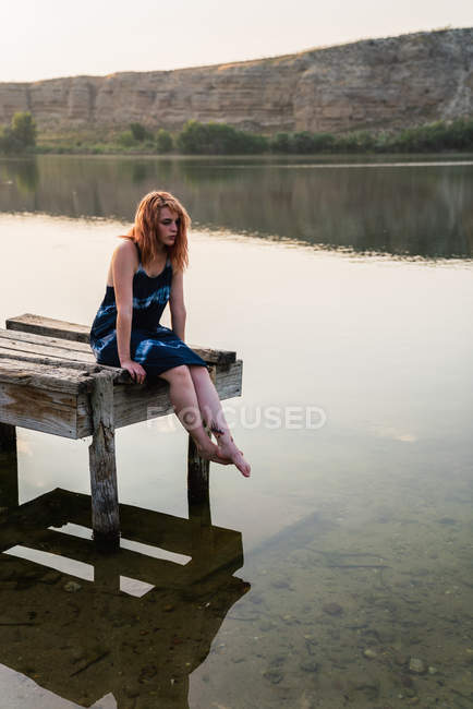 Чувственная молодая женщина, сидящая на пирсе в озере на природе — стоковое фото