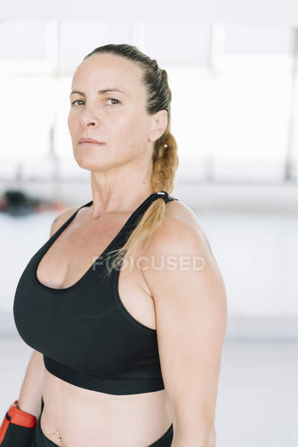Femme confiante en vêtements de sport debout sur fond flou de gymnase et en regardant la caméra — Photo de stock