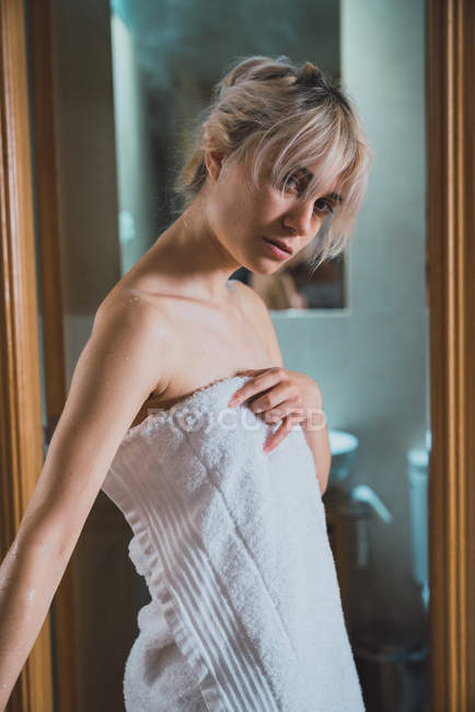 Блондинка, завернутая в белое полотенце, смотрит в камеру в дверях ванной комнаты — стоковое фото