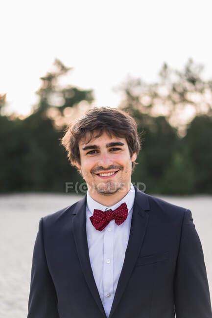 Homem adulto vestindo elegante terno preto com gravata borboleta e em pé na praia com as mãos nos bolsos olhando para a câmera — Fotografia de Stock