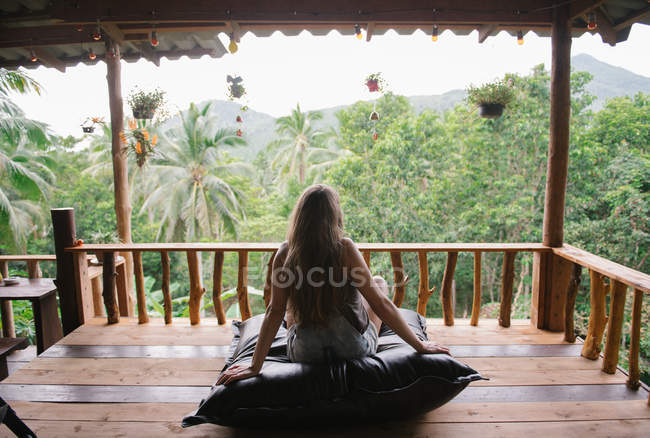 Вид сзади женщины, сидящей и смотрящей на пальмы на террасе в тропическом лесу — стоковое фото