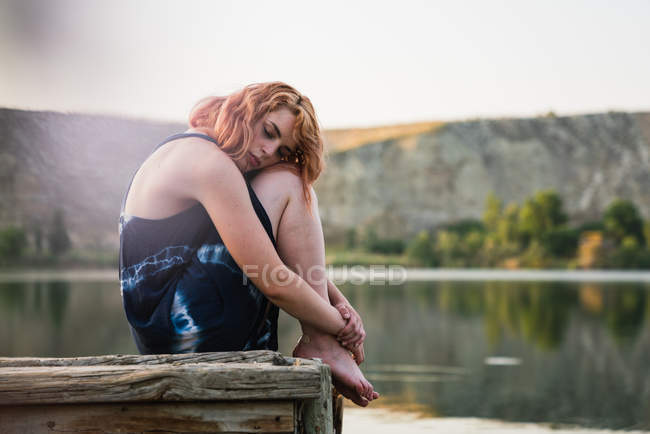 Sensual jovem mulher sentada no cais abraçando joelhos no lago na natureza — Fotografia de Stock