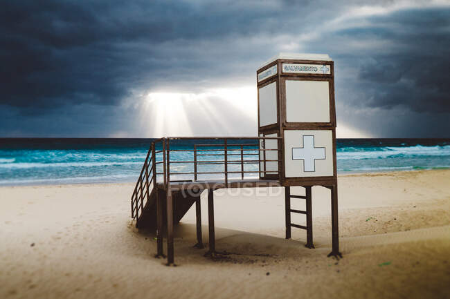 Маленькая белая будка на деревянной платформе с поперечным знаком на стенах, расположенных на прекрасном песчаном берегу океана в пасмурный день с приближающимся штормом на Фуэртевентуре, Канарские острова — стоковое фото