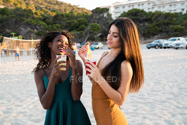 Молодые подруги отдыхают на пляже с напитками в чашках и смеются во время чата на закате — стоковое фото