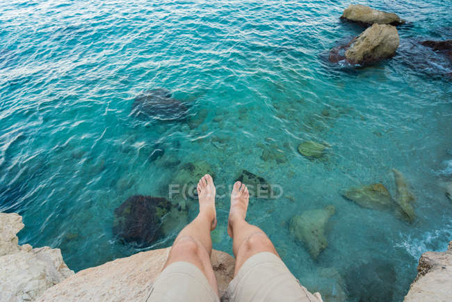 Legs of man sitting on cliff above turquoise water — Stock Photo