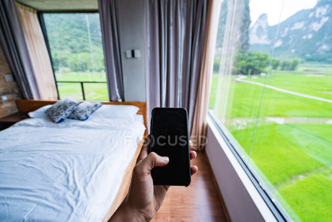 Cortar mão segurando smartphone em um quarto elegante com vista para as montanhas chinesas — Fotografia de Stock